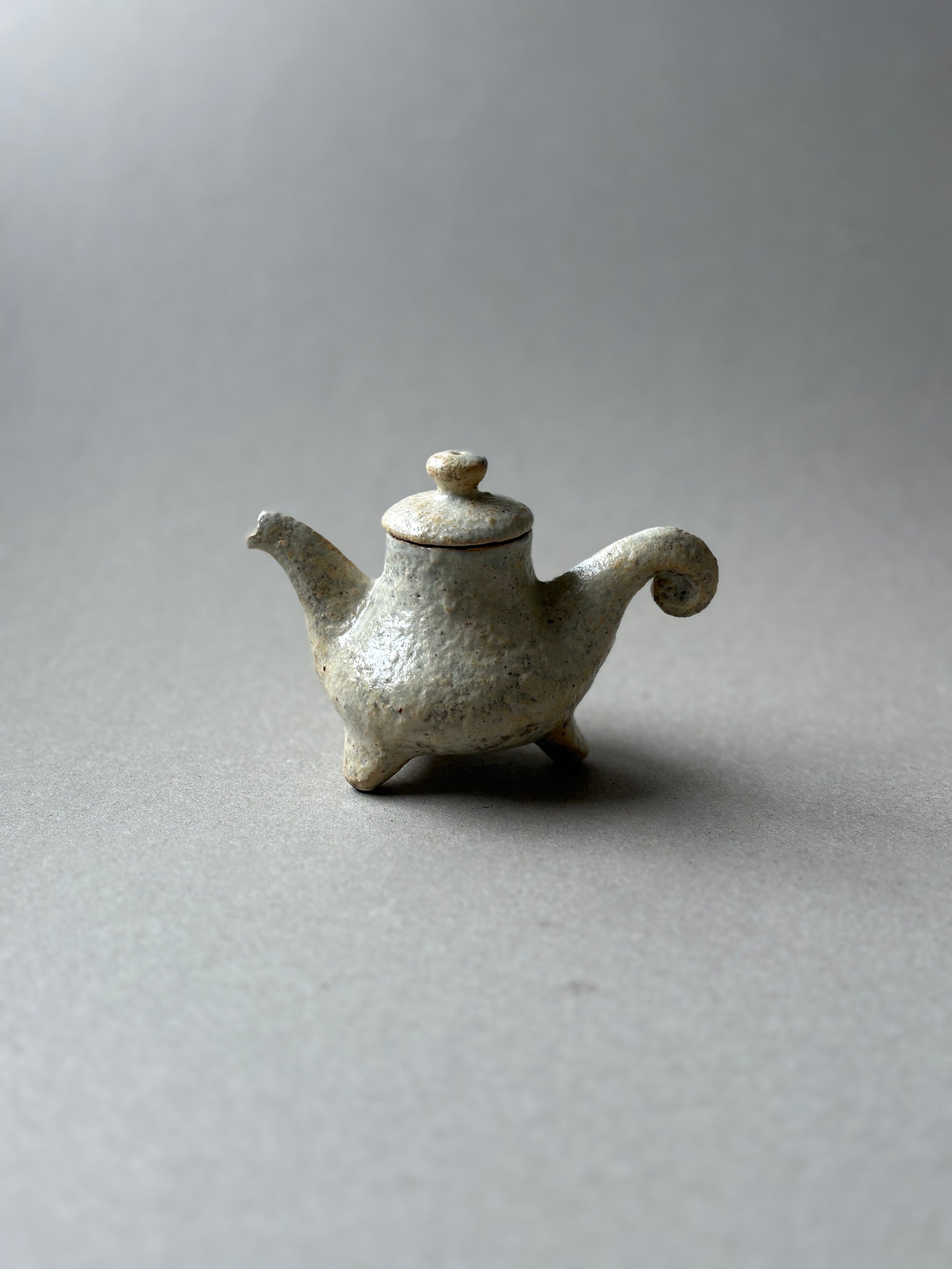 A small handmade ceramic teapot with a textured, speckled off-white glaze, featuring a curved spout, a lid with a rounded knob, and three short legs, set against a neutral gray background.