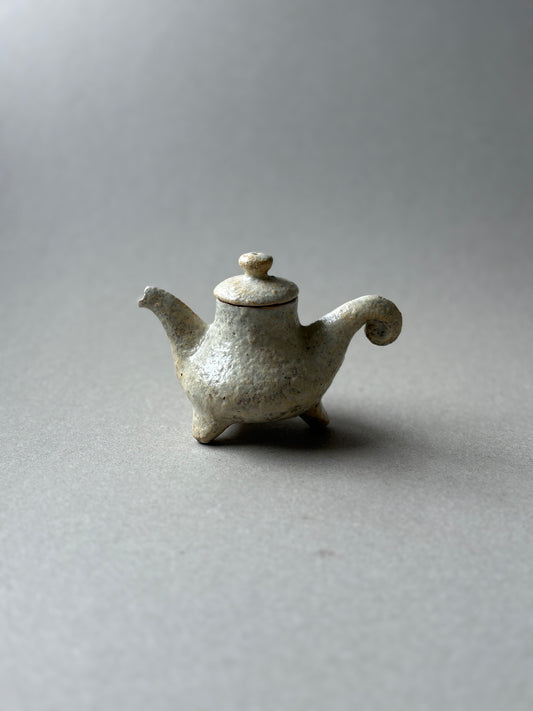 A small handmade ceramic teapot with a textured, speckled off-white glaze, featuring a curved spout, a lid with a rounded knob, and three short legs, set against a neutral gray background.
